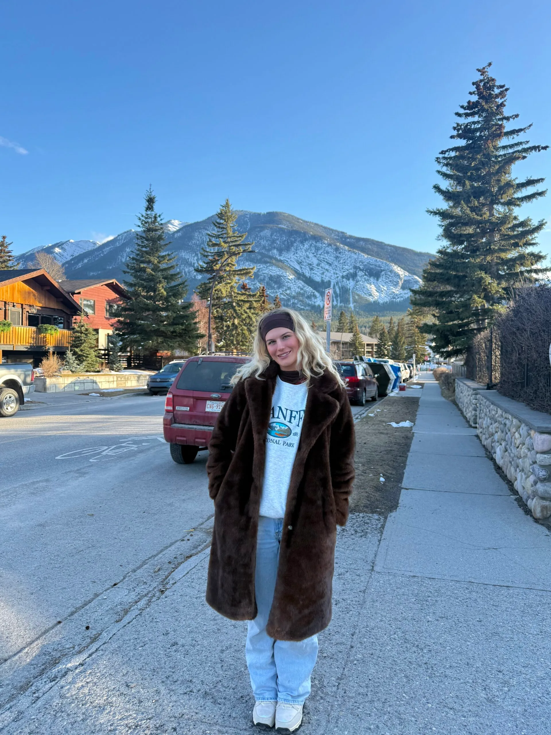 Woman wearing jumper and pants and standing infront of mountain, smiling at camera.