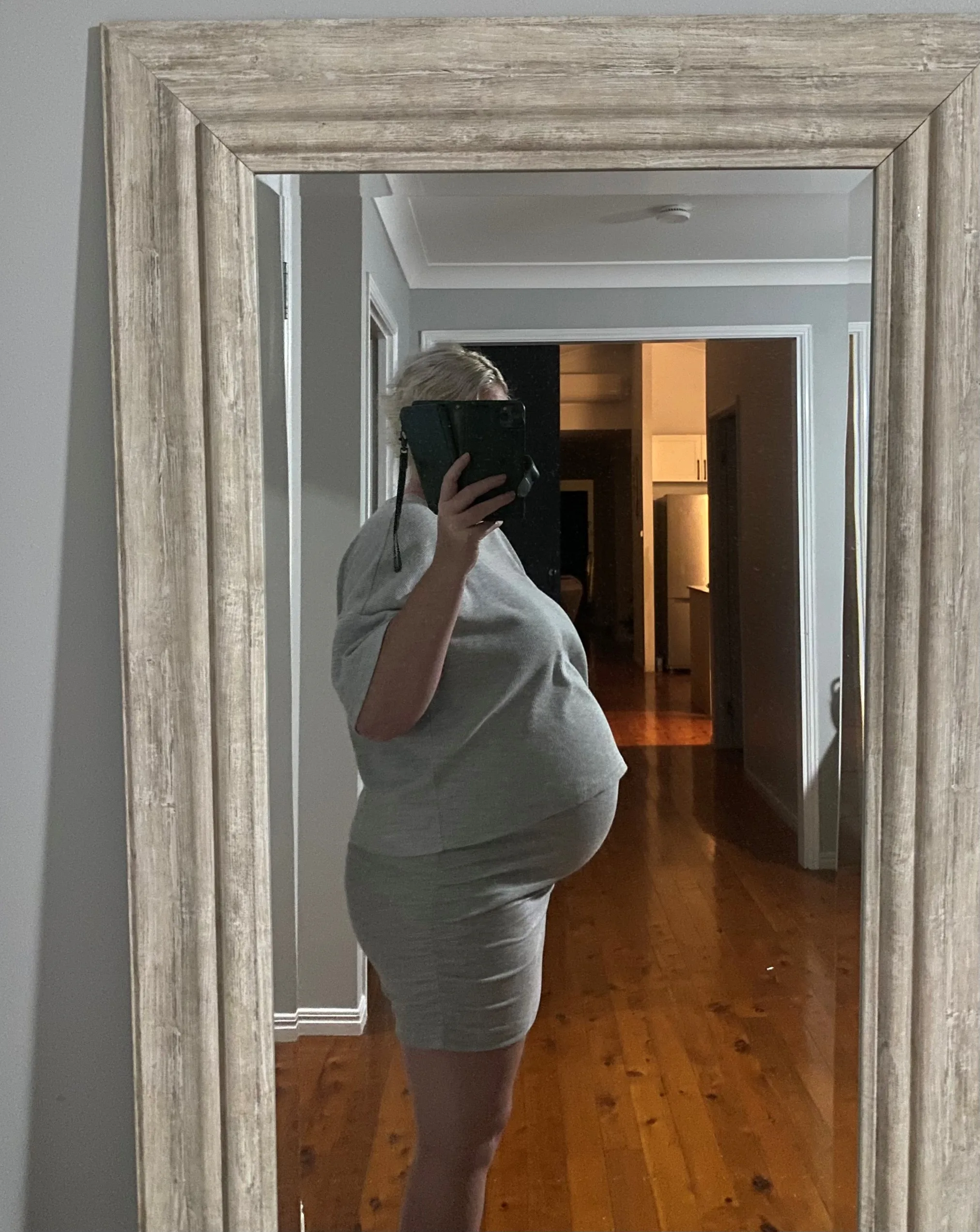 Pregnant woman wearing grey top and skirt and standing infront of mirror.