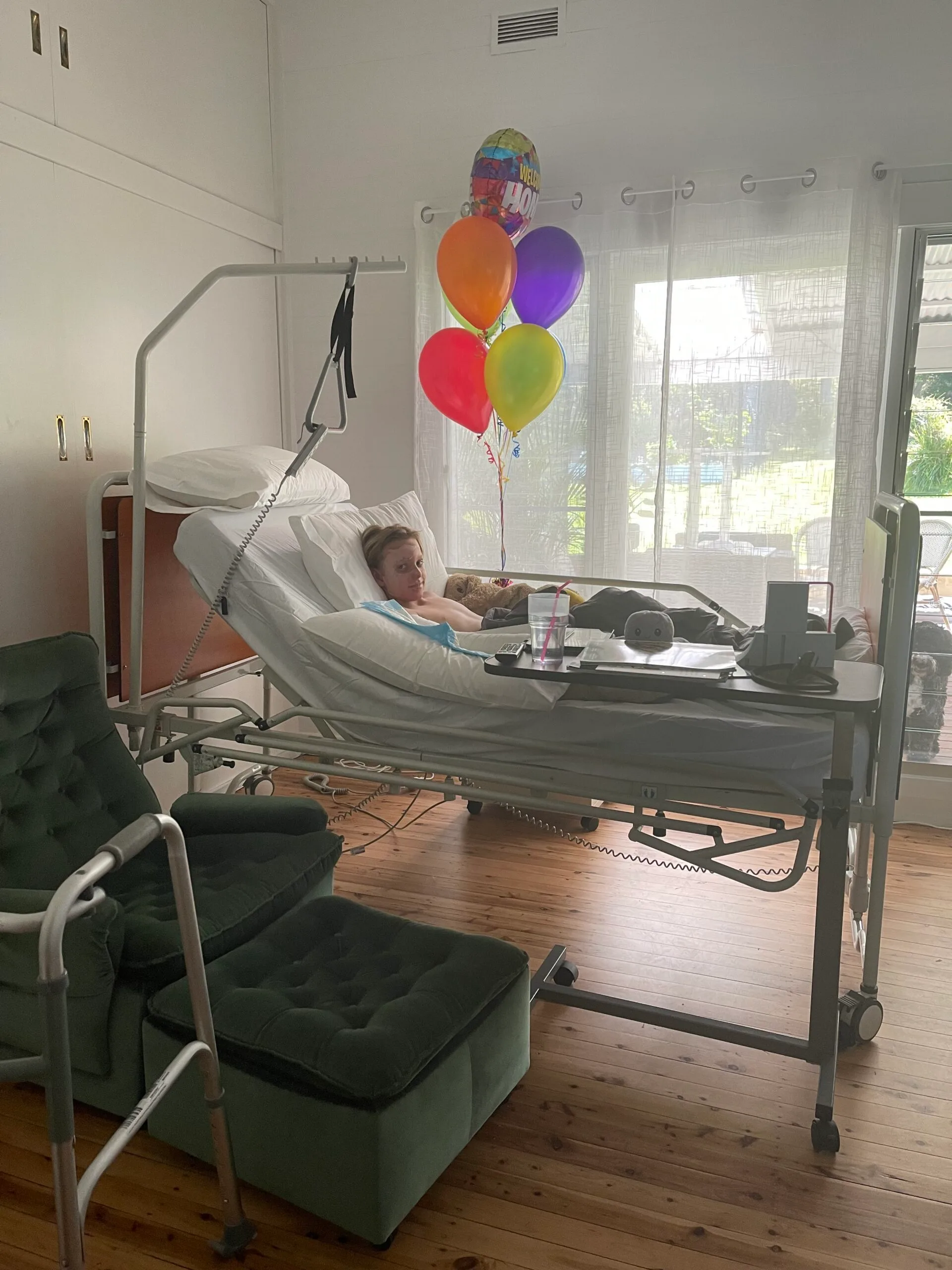 Young boy in hospital bed resting next to colourful balloons.