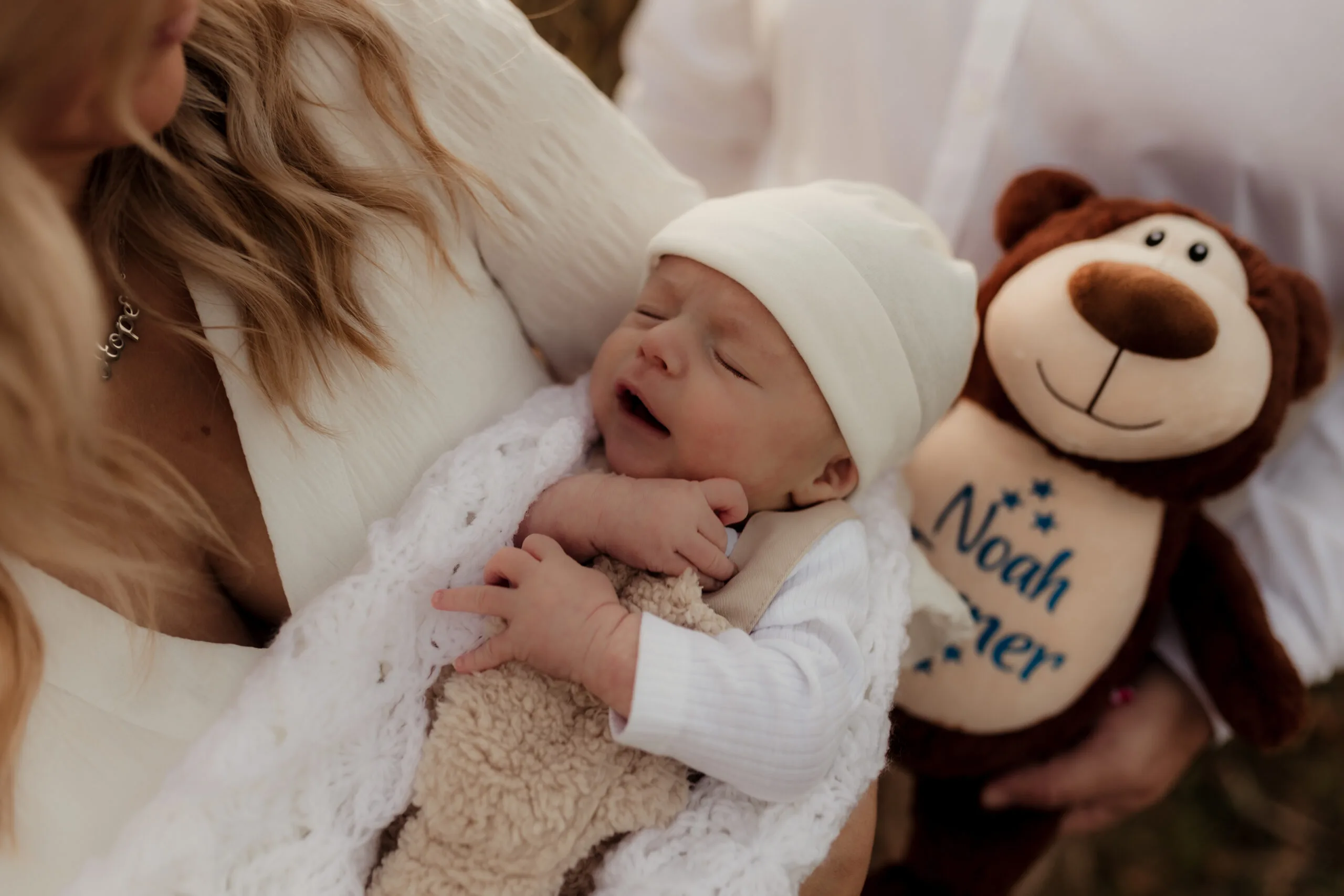 Mum holding newborn baby wearing white beanie and clutching onto soft toy.