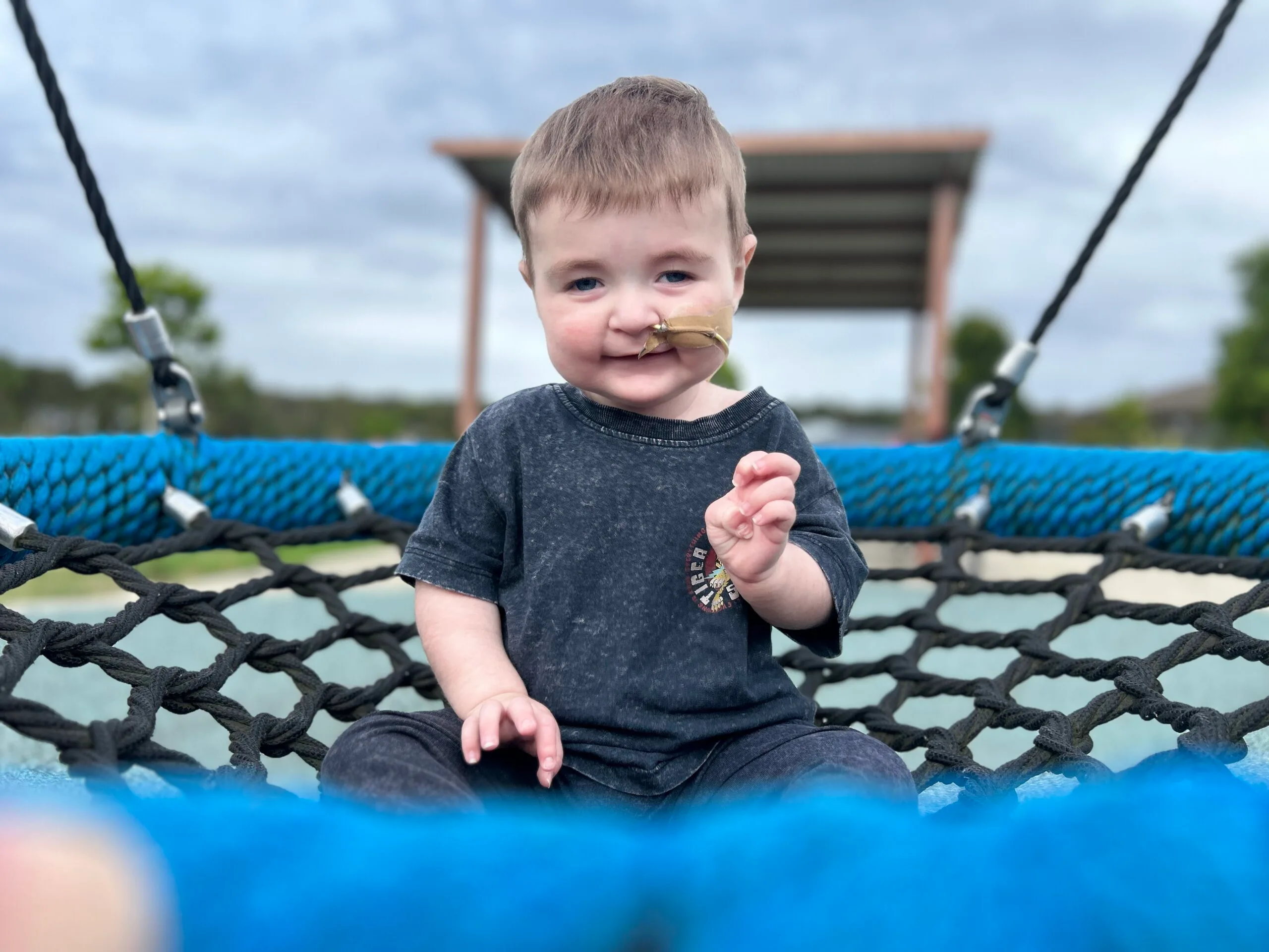 Toddler with feeding tube, wearing black shirt and pants and sitting on swing, smiling at camera.