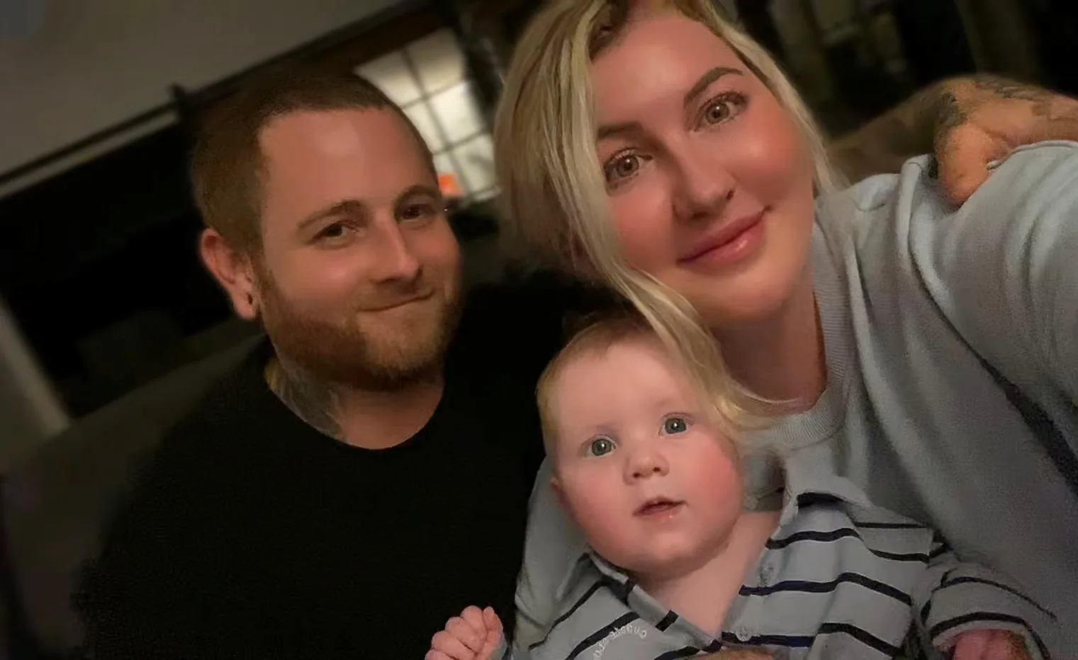 Man wearing black shirt, next to blonde woman holding baby and all smiling at camera.