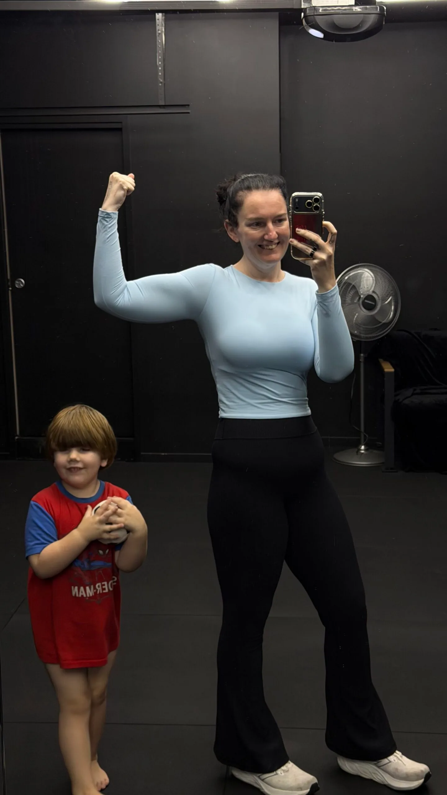 Woman in gym wearing blue top and leggings with toddler next to her. Both smiling at camera.