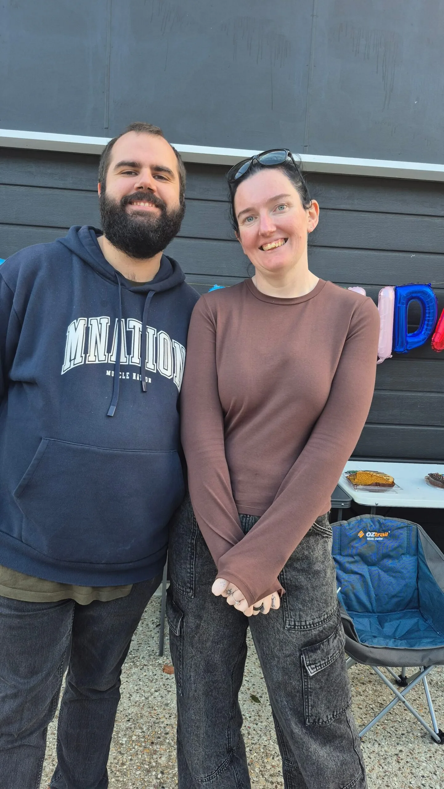 Man wearing blue jumper and pants next to woman wearing brown long sleeve top and smiling at camera.