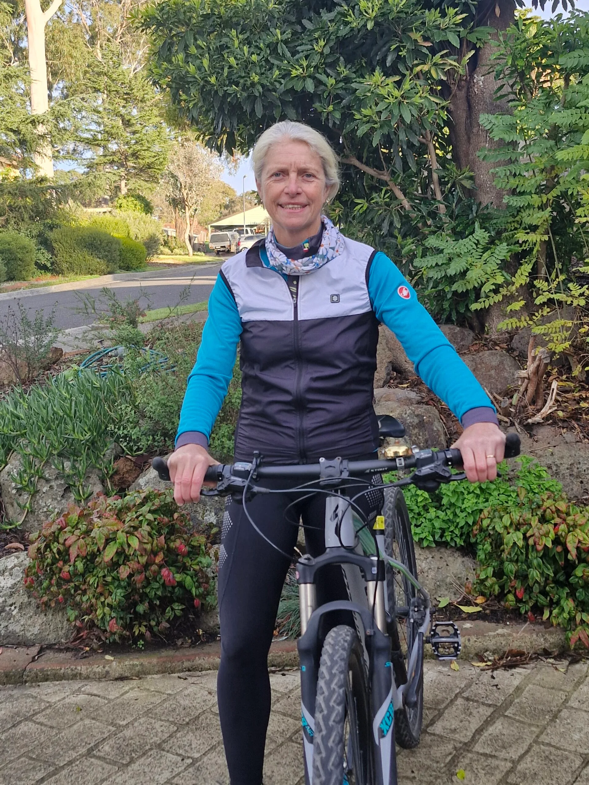 Woman wearing blue and white jacket and black leggings, standing with black bike and smiling at camera.
