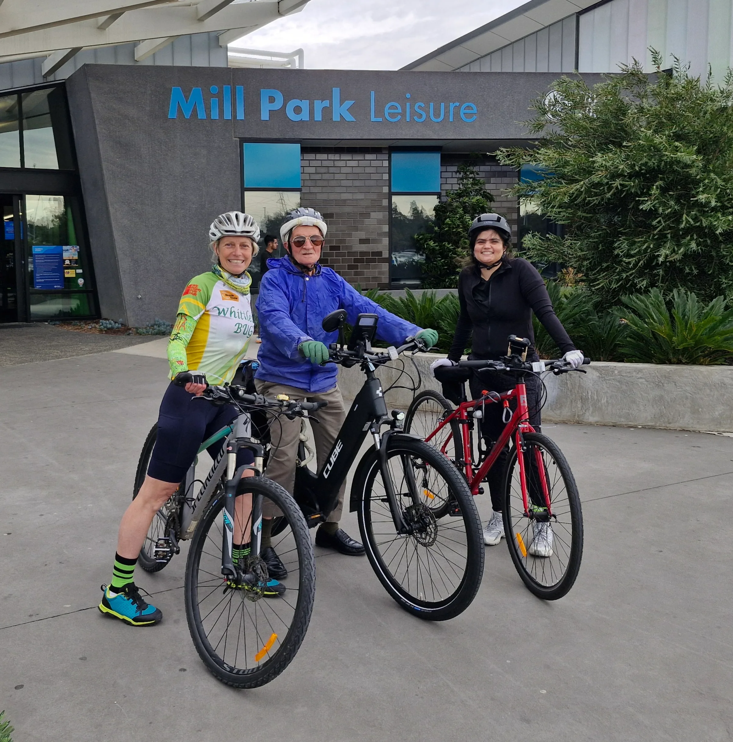 Two women on black bikes alongside a man on a black bike. All wearing helmets and smiling at camera.
