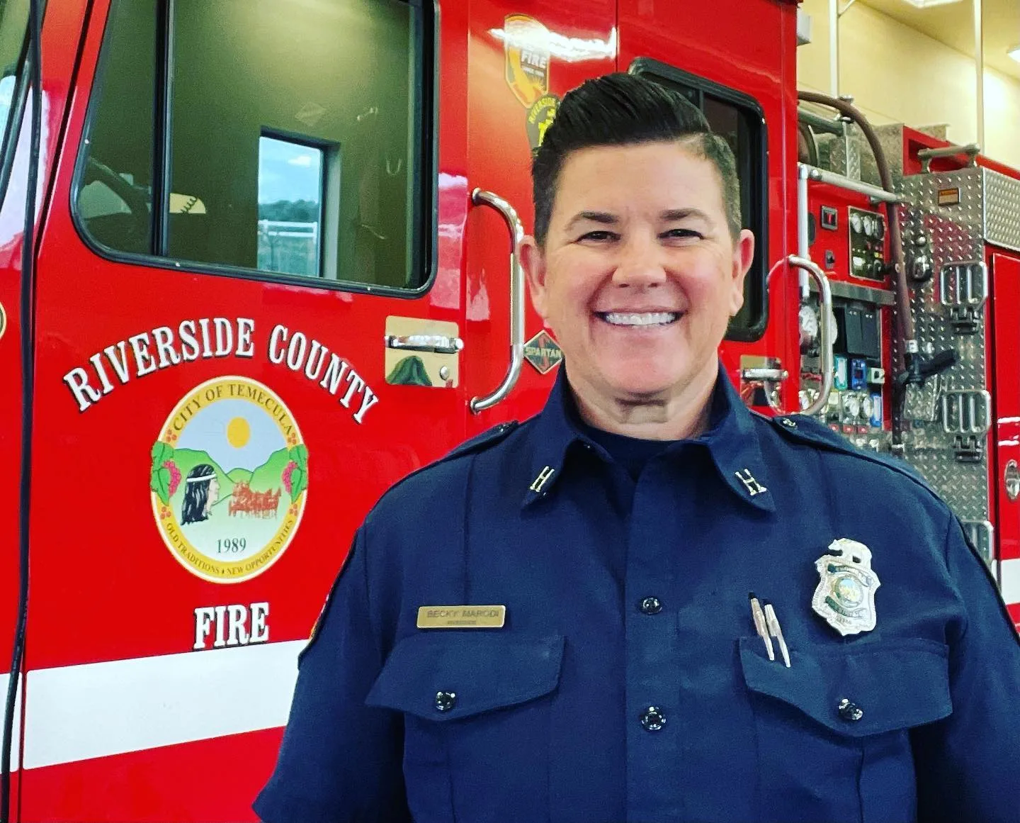 Woman wearing firefighter uniform smiling infront of firetruck.