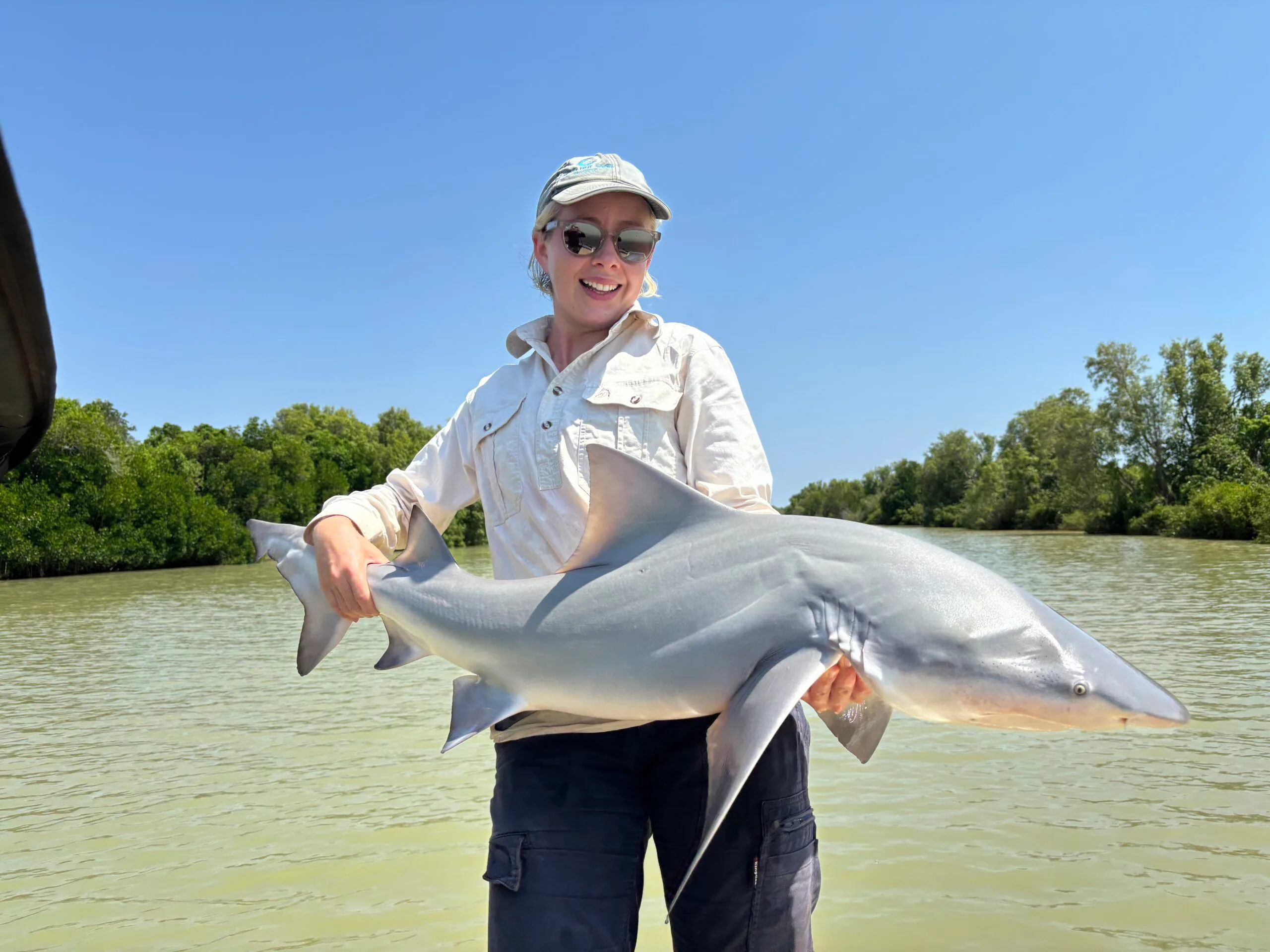 Woman wearing white button up top and holding up a shark in the water.