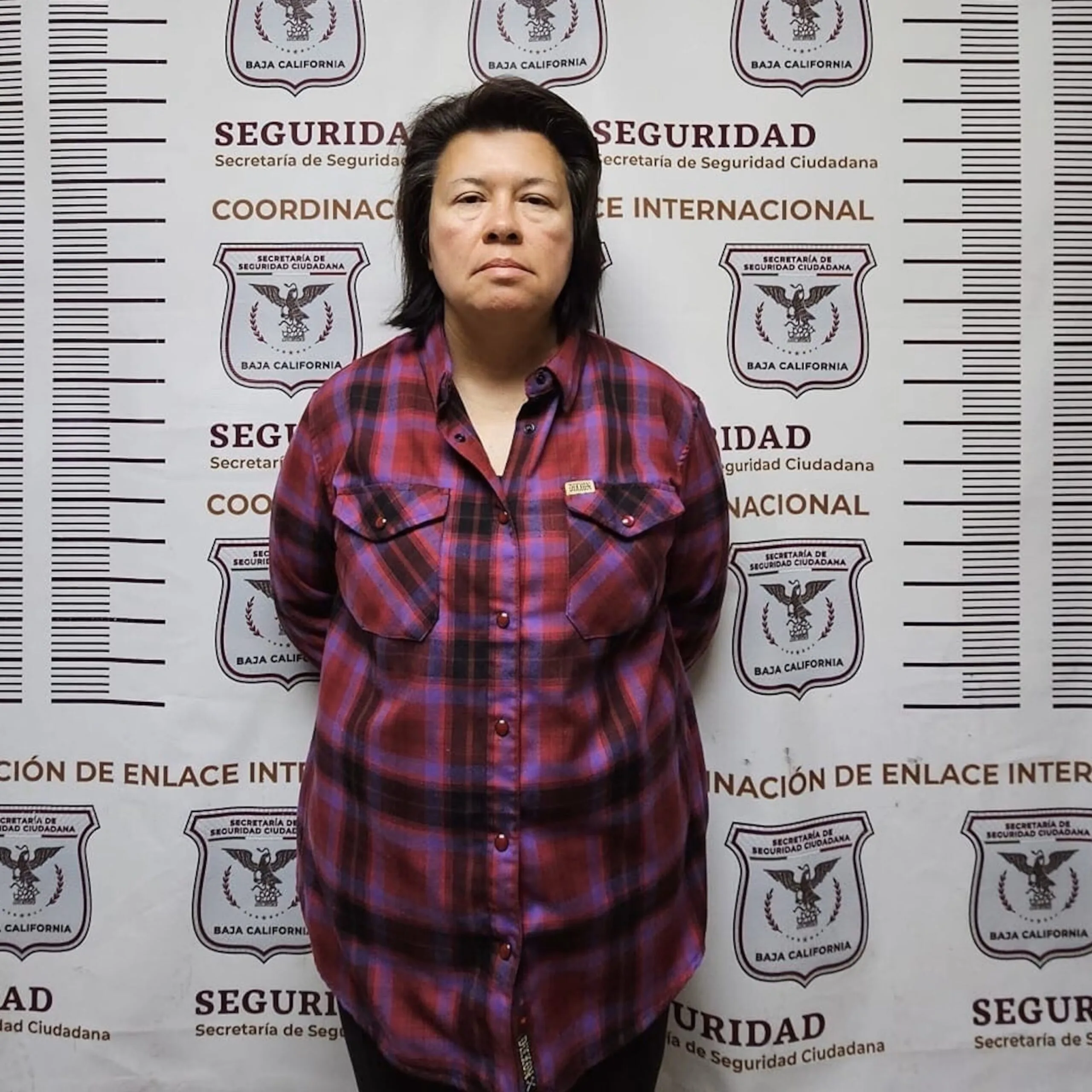 Mugshot of woman with short hair, wearing checkered shirt and staring at camera.