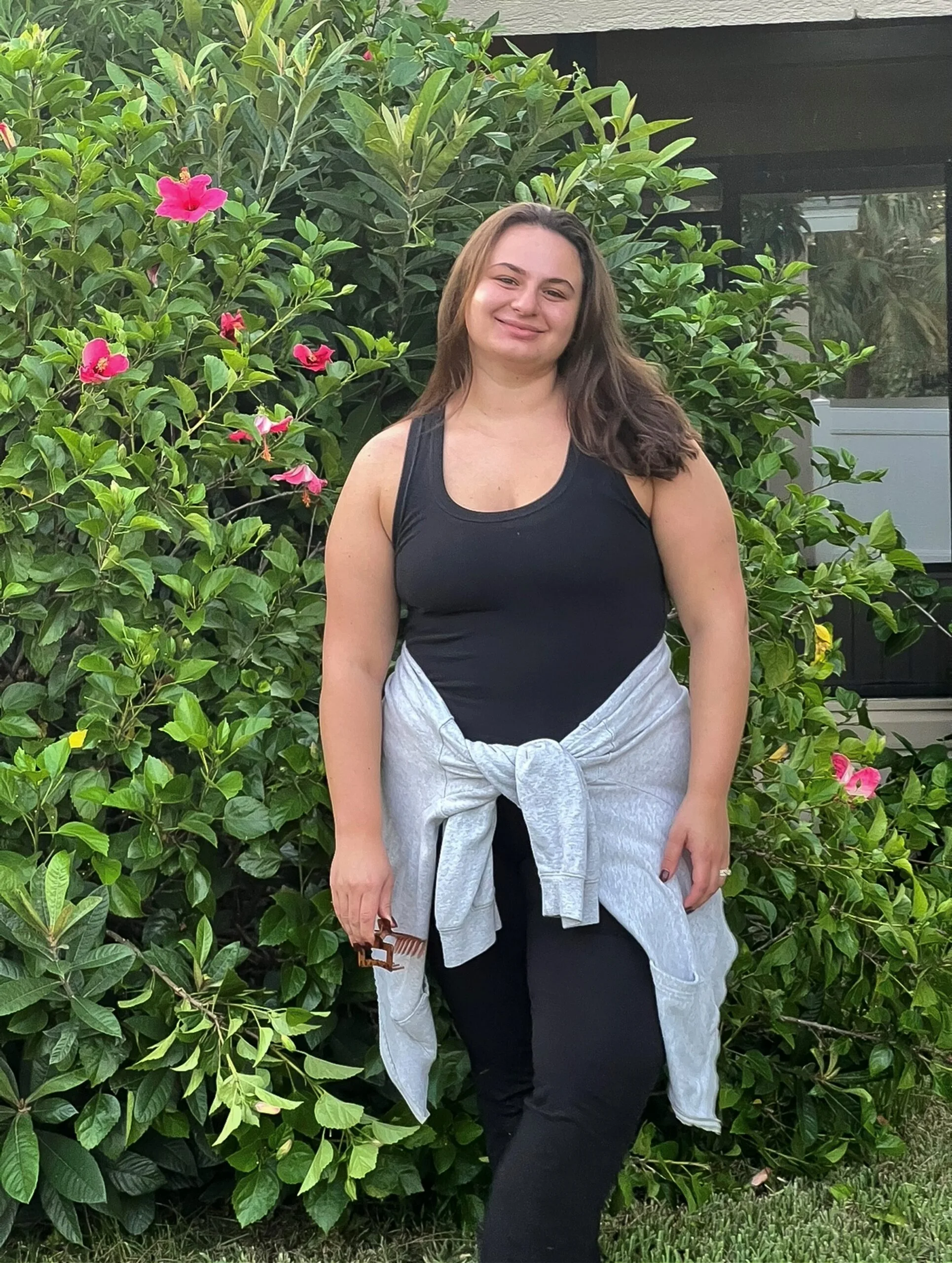Young woman, overweight, standing infront of bush, smiling.
