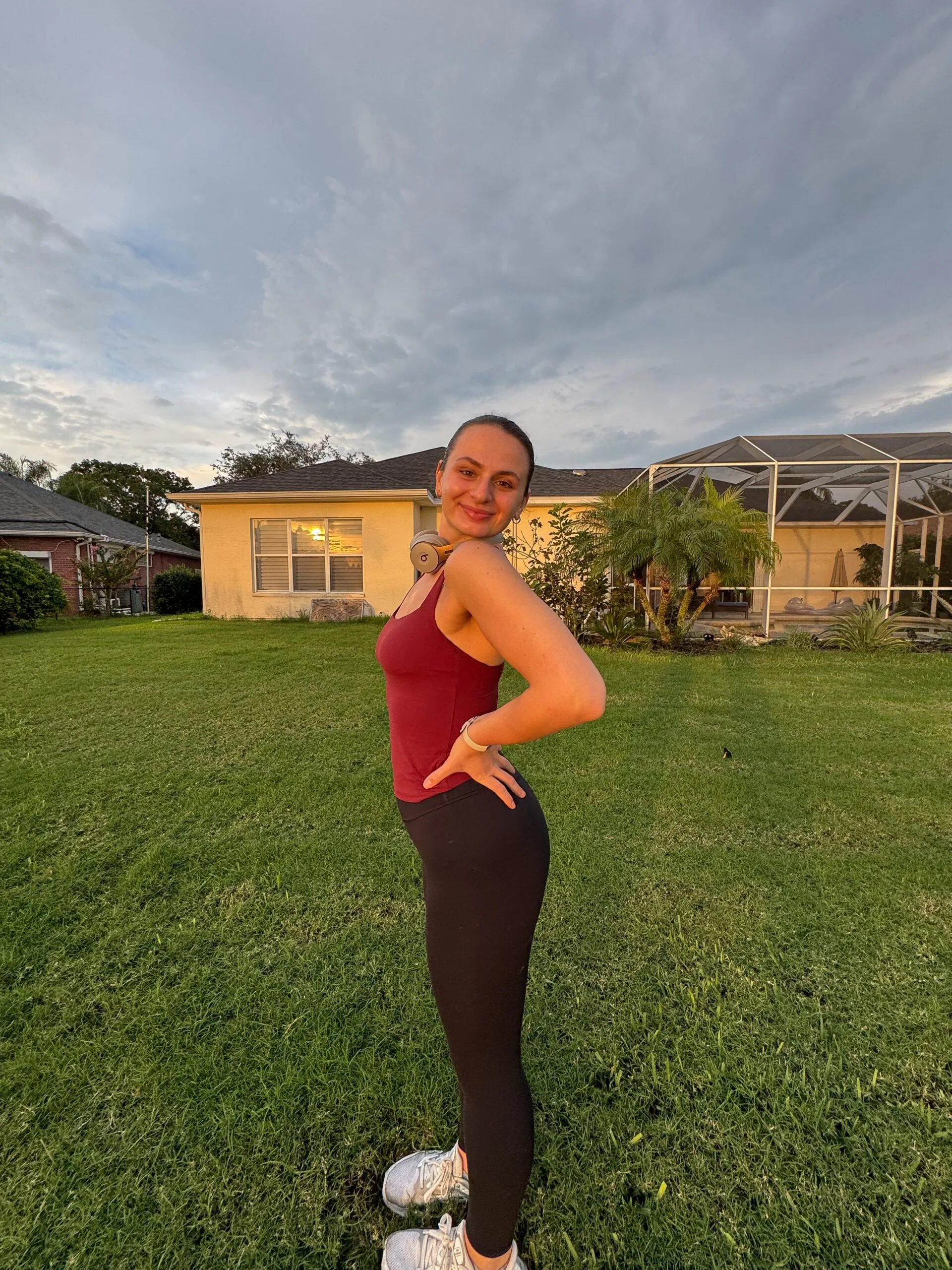 Woman wearing red top and leggings, standing on lawn and smiling at camera.
