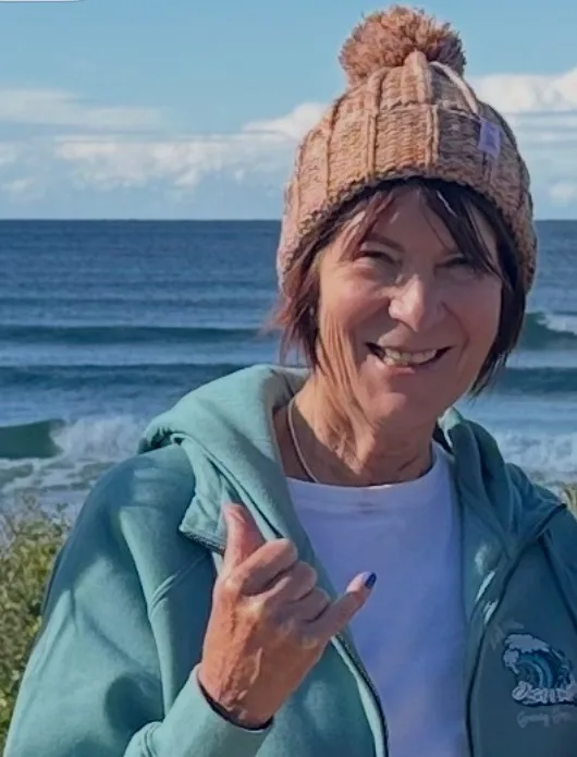 Woman with short red hair, wearing beanie and jacket, standing infront of ocean and smiling at camera.