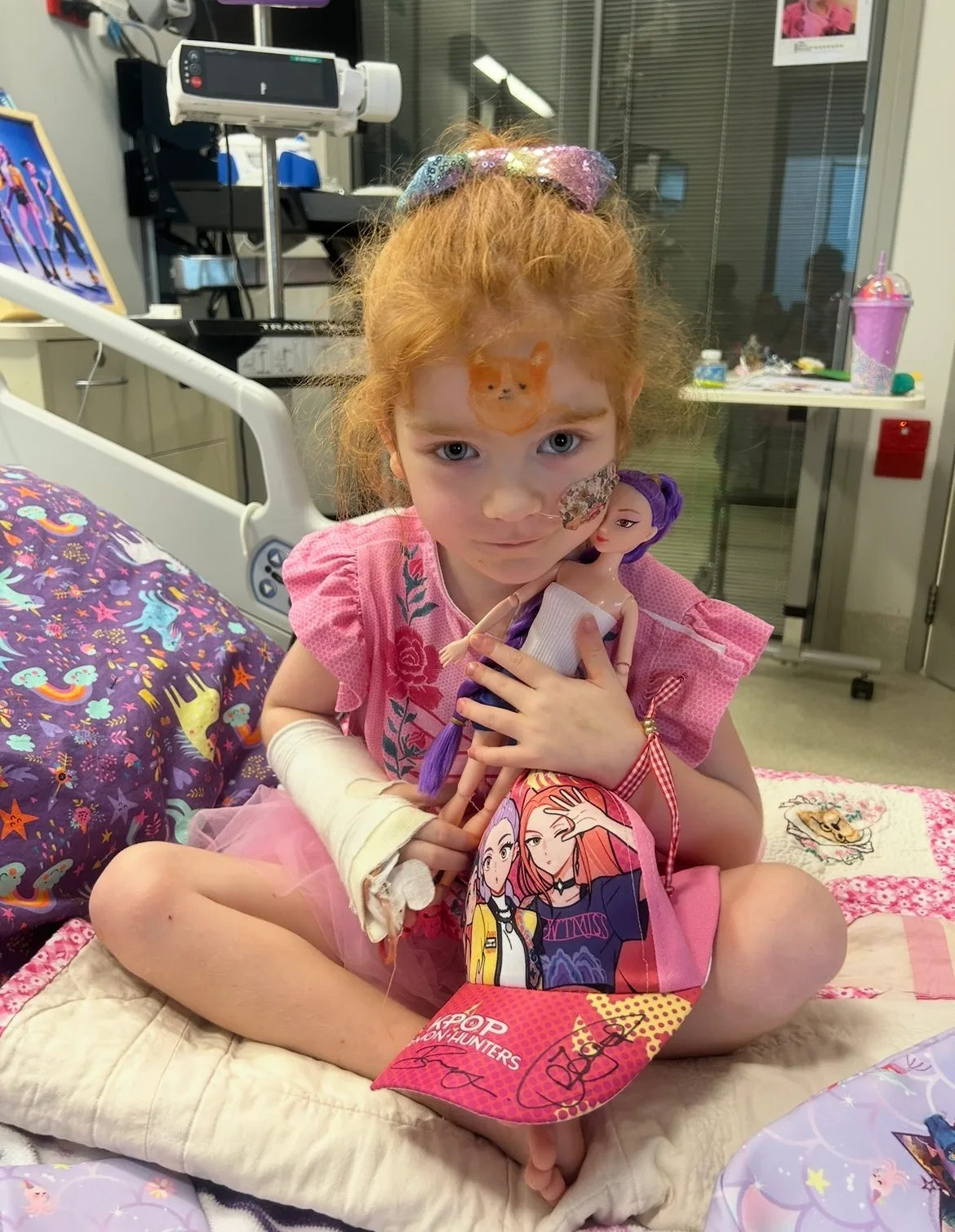 Young girl with strawberry blonde hair, wearing pink dress and holding barbie doll sitting on hospital.