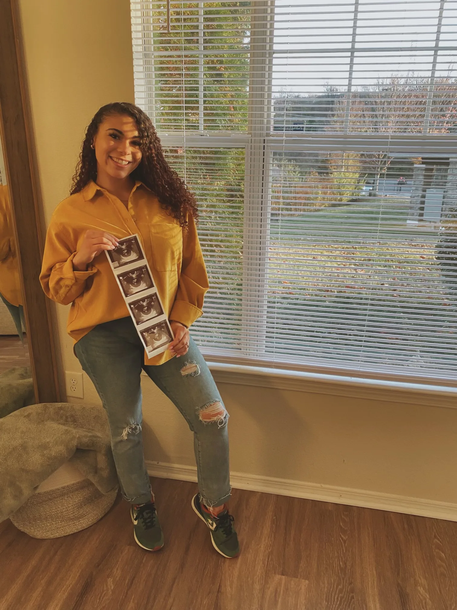 Woman wearing yellow shirt holding up strip of ultrasound photos of baby while smiling at camera.