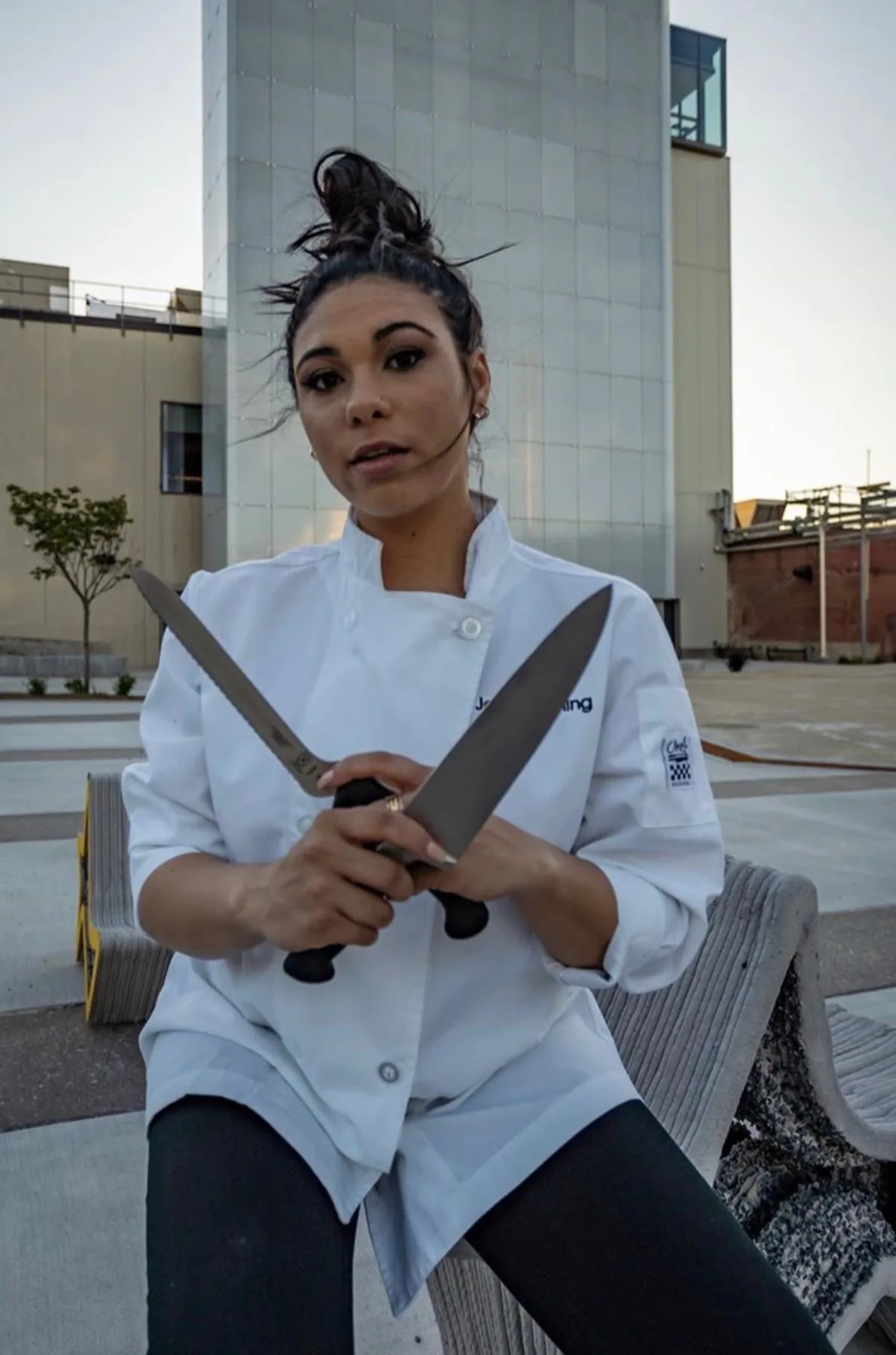 Woman with dark hair, wearing chef attire and holding up knives, staring at camera.