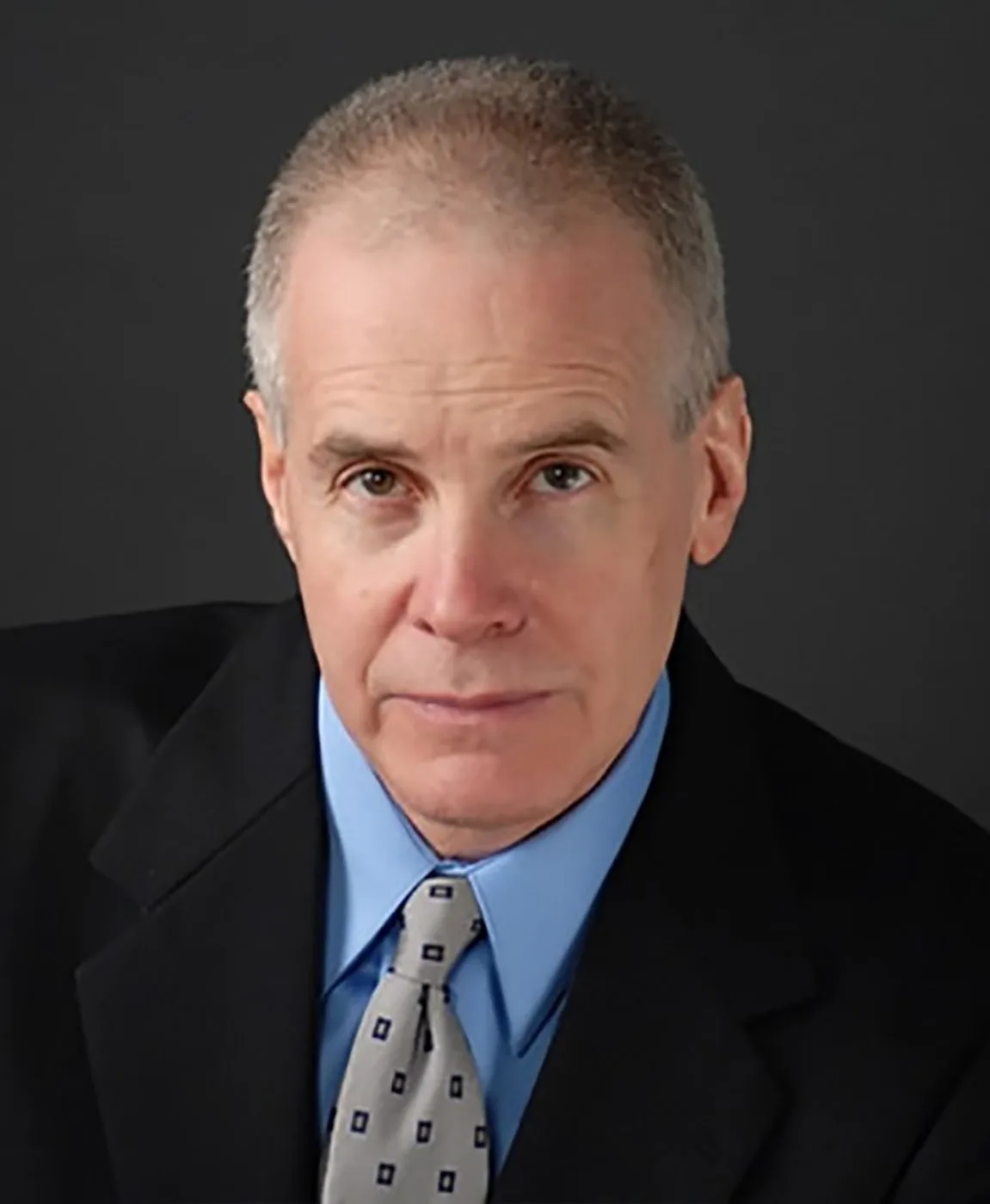 Professional photo of man with grey hair, wearing blue shirt and black suit, staring at camera.