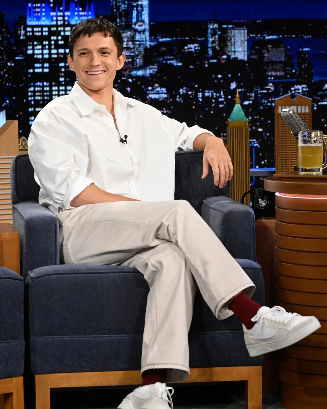 celebrity gemini tom holland smiling on a talk show couch wearing a white jumper and beige trousers
