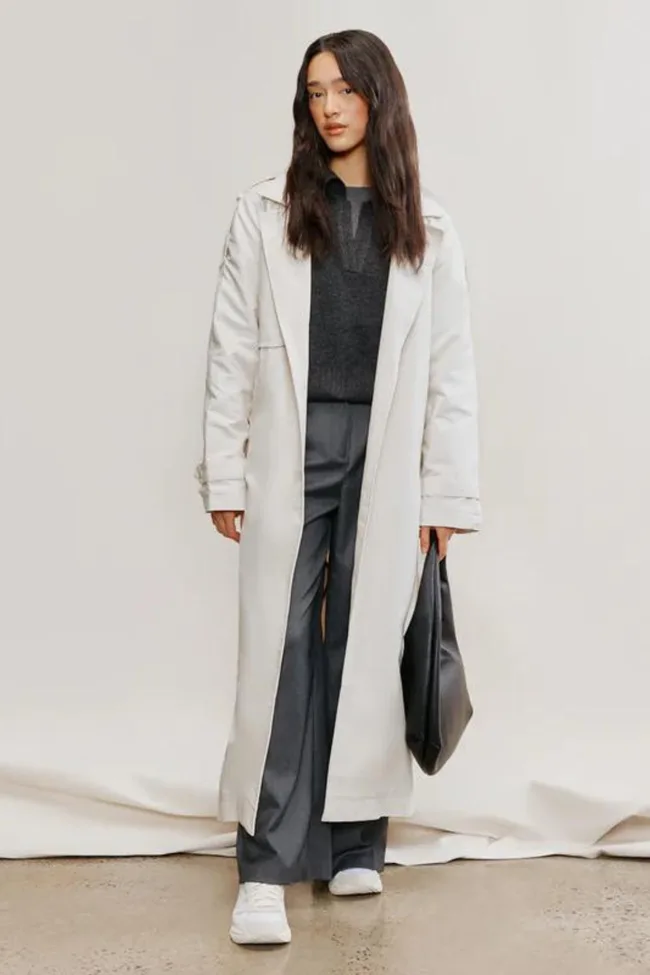 A person in a long white coat over a dark sweater and gray pants, holding a black bag, standing against a plain background.
