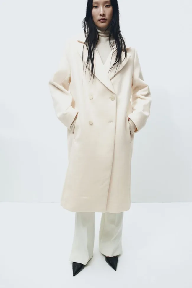Model wearing a long cream overcoat with hands in pockets, white pants, and black pointed shoes.