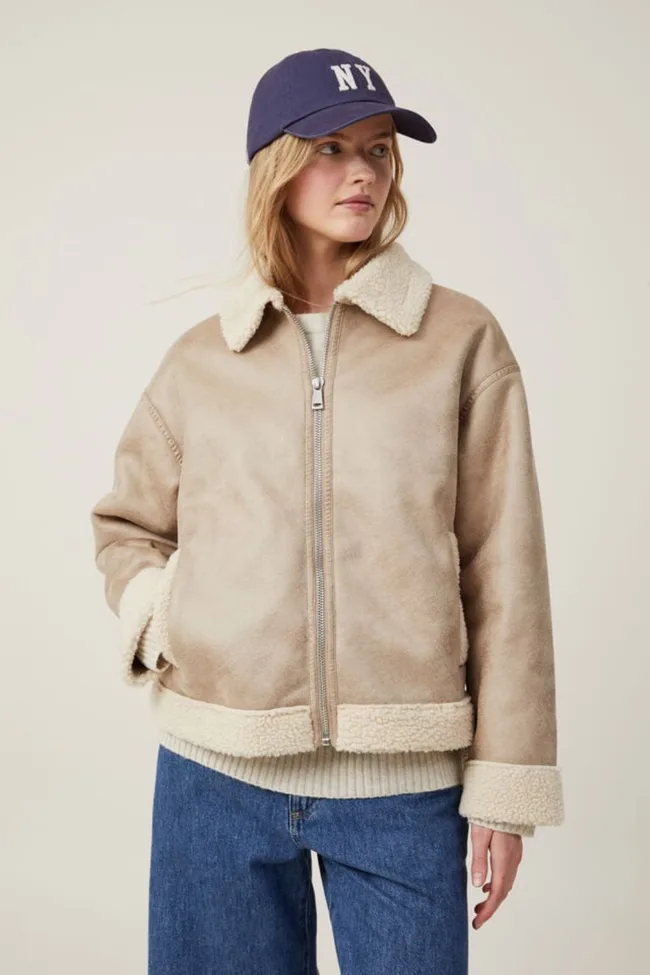 Young woman in a shearling jacket and NY cap, looking sideways, with hands in pockets.