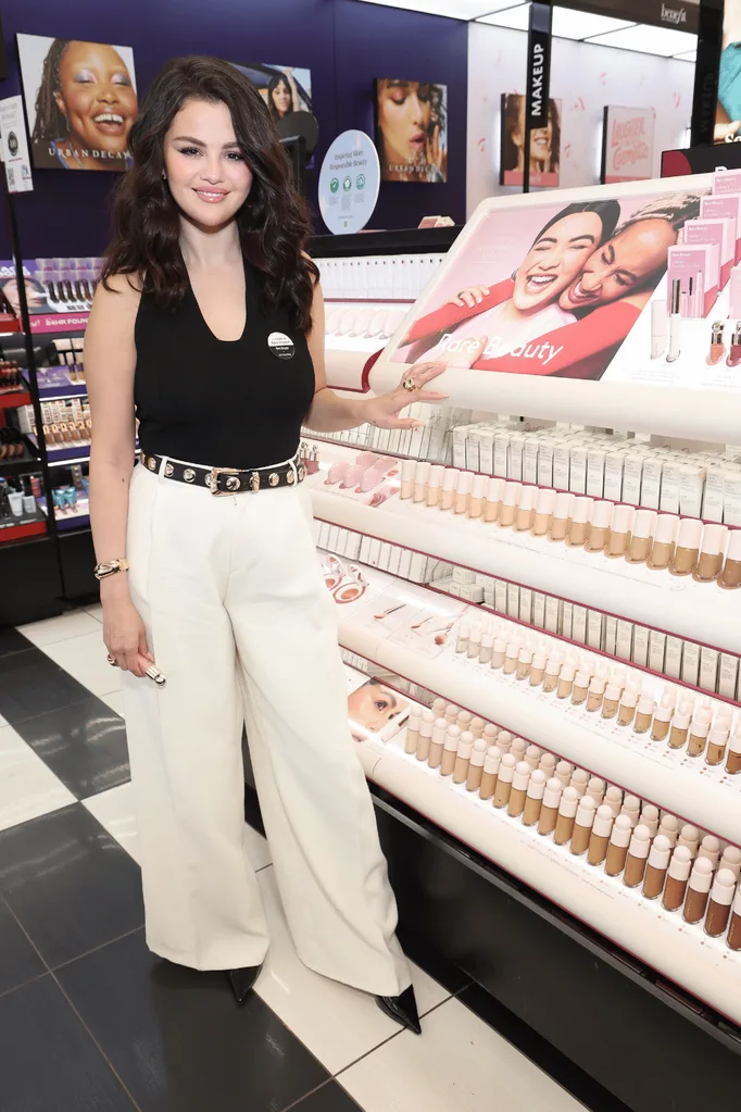 Selena Gomez standing next to a Rare Beauty Sephora display stand.