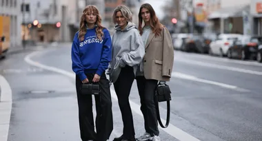 Three women standing on a city street, wearing casual streetwear and holding handbags.