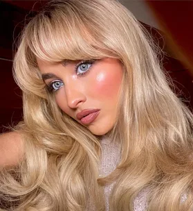 Blonde woman with long hair, bangs, and blue eyes, wearing shimmering makeup and a sparkly top.