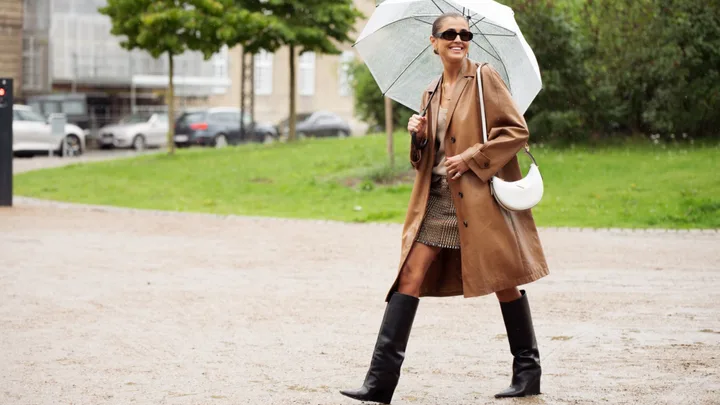 A woman in a brown coat and sunglasses walks with an umbrella on a rainy day.