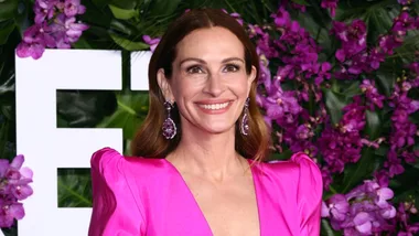 A person in a bright pink dress and earrings smiles against a backdrop of purple flowers and foliage.