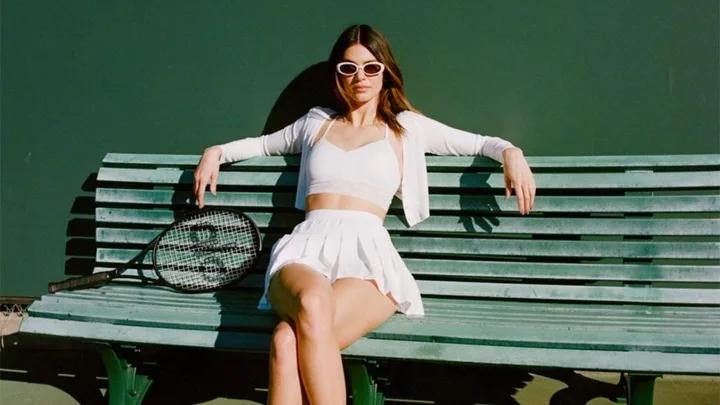 Woman in white outfit and sunglasses sits on a bench with a tennis racket beside her.