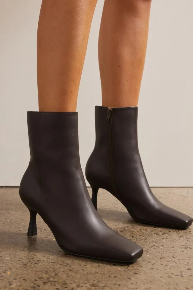 Black ankle boots with thin heels and side zips on feet standing on a textured floor.