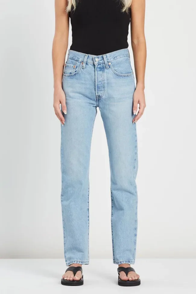 Woman wearing a black top, light blue jeans, and black sandals, standing against a plain background.