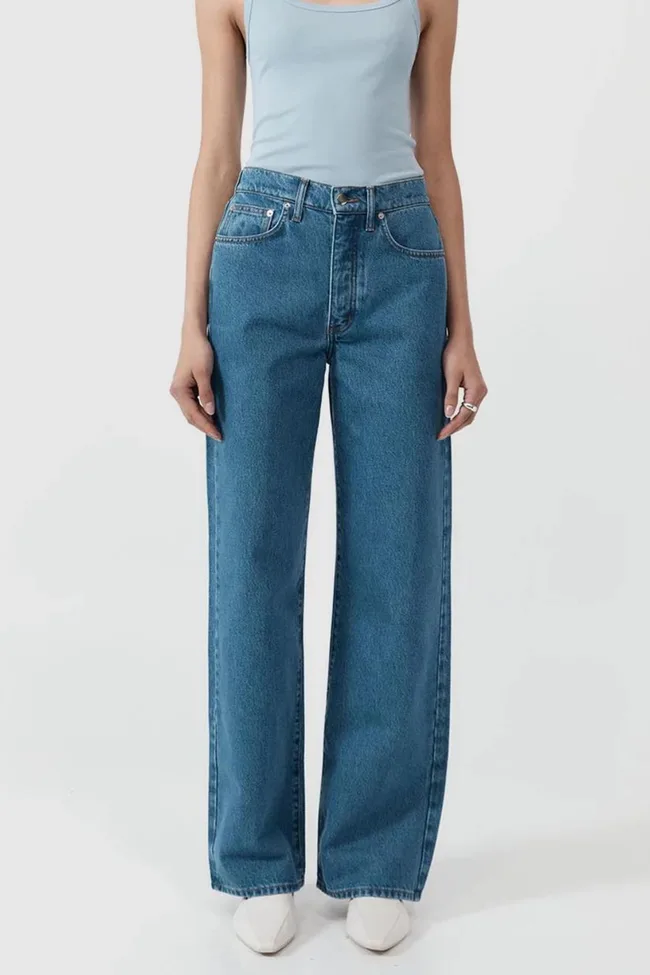 Person wearing light blue tank top and high-waisted blue jeans with wide legs; white shoes visible; hands at sides.