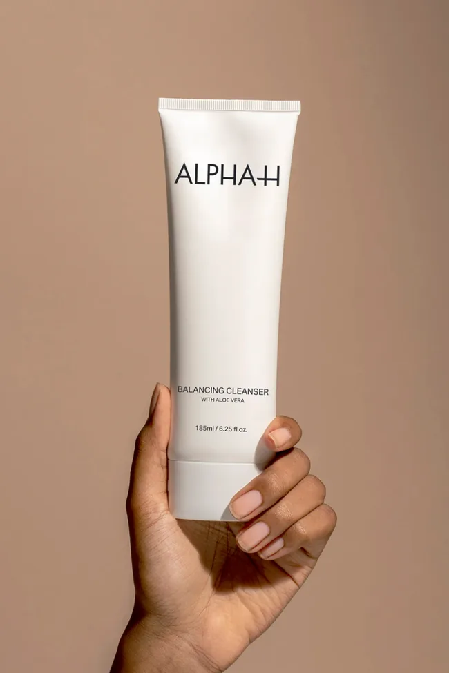 Hand holding a tube of Alpha-H Balancing Cleanser with Aloe Vera, against a beige background.