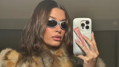 Person in sunglasses takes a mirror selfie wearing a fur coat, holding a phone and a tube of beauty product.