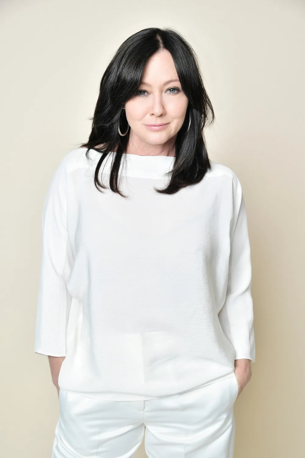 Portrait of Shannen Doherty wearing a white shirt and pants.