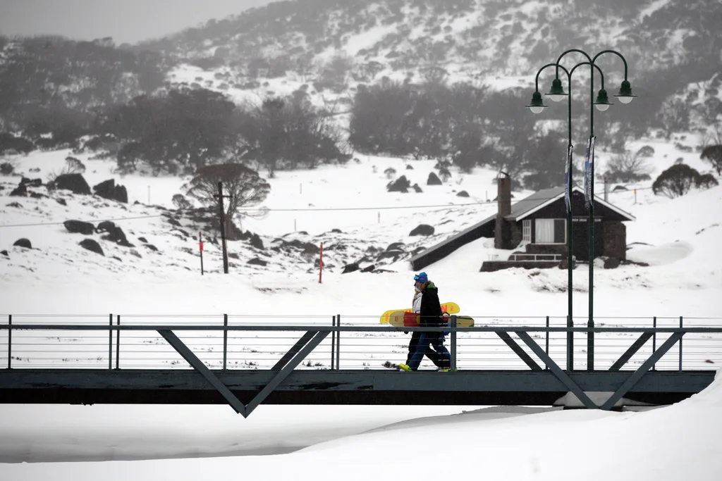 Two snowboarders walking across a bridge surrounded by snow. 