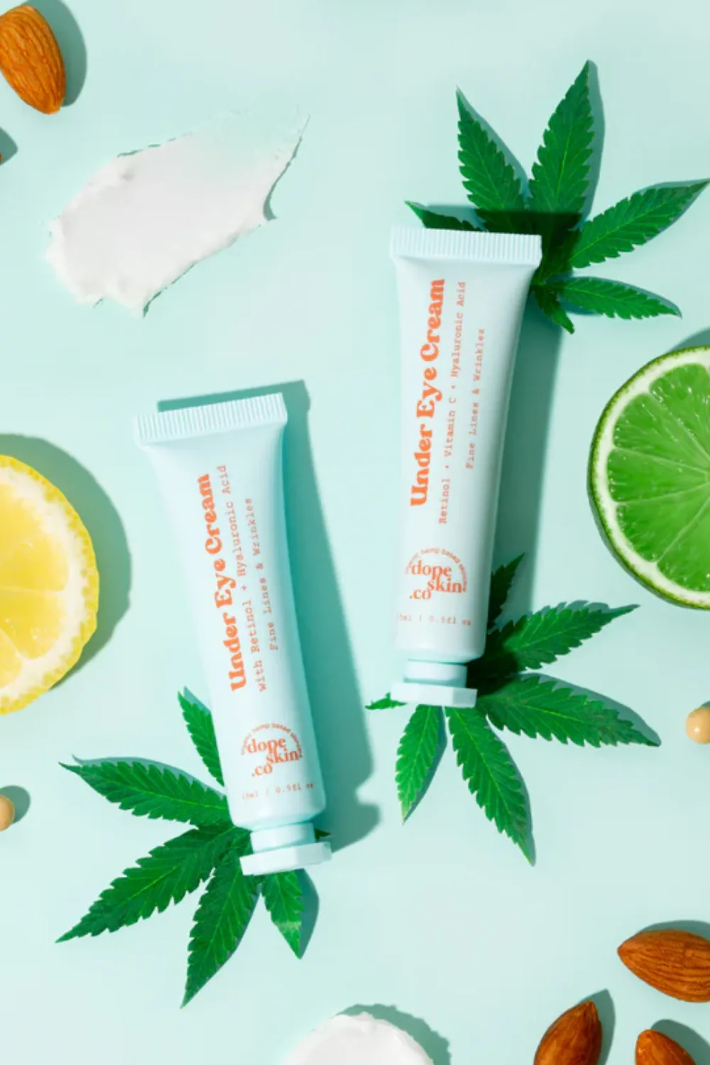 Dope Skin Co's Under eye cream on a blue background surrounded by hemp leaves, limes, and lemons.