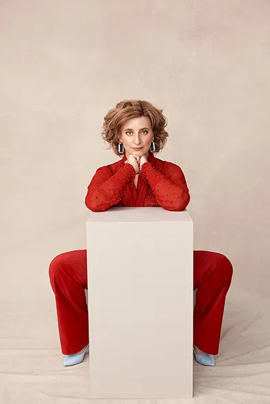 Felicty Ward wears a red suit and leans on a big box