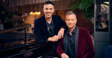 Two men in velvet jackets pose by a piano in a warmly lit room.