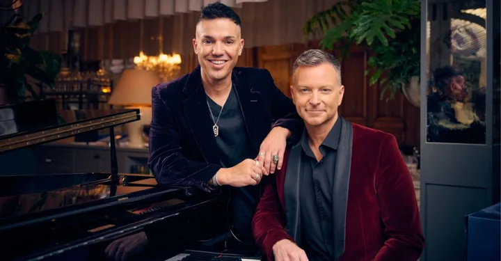 Two men in velvet jackets pose by a piano in a warmly lit room.