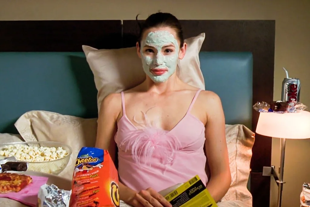 Girl in pink nightgown watching a movie in bed with a face mask and bowl of popcorn