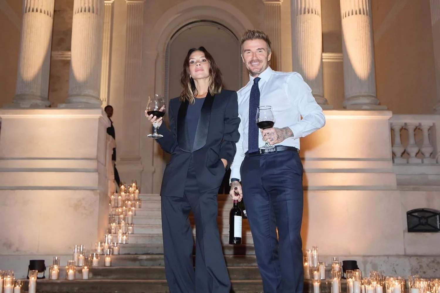 Victoria Beckham and David Beckham stand on a staircase each holding a glass of red wine