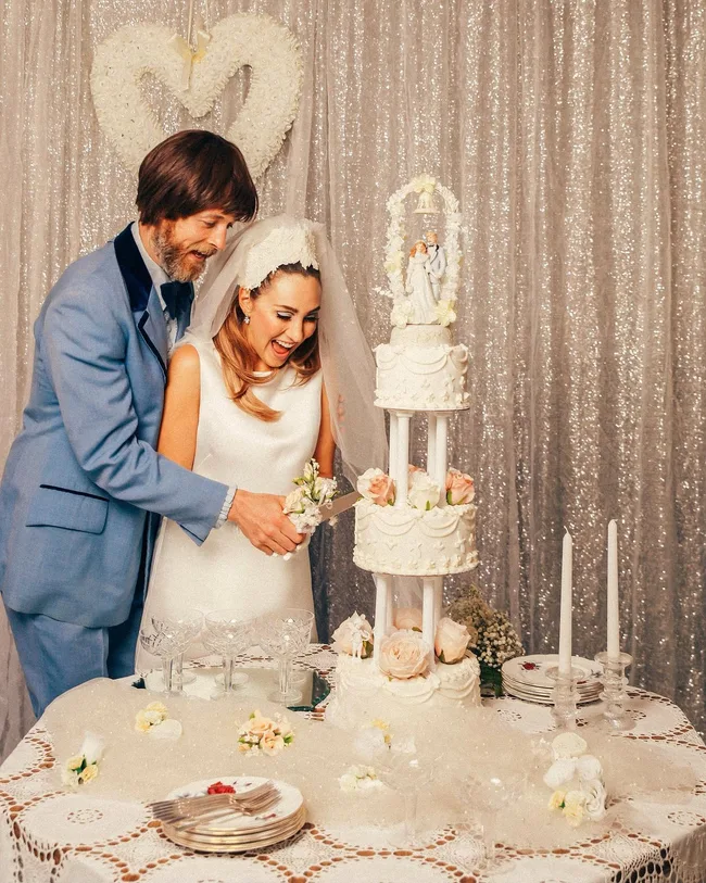 Hamish Blake and Zoe Foster Blake cutting a tiered wedding cake while dressed in 1960s attire.
