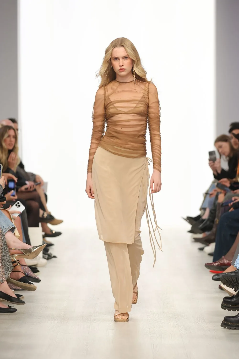 Model walks a runway way in a sheer beige top and pants
