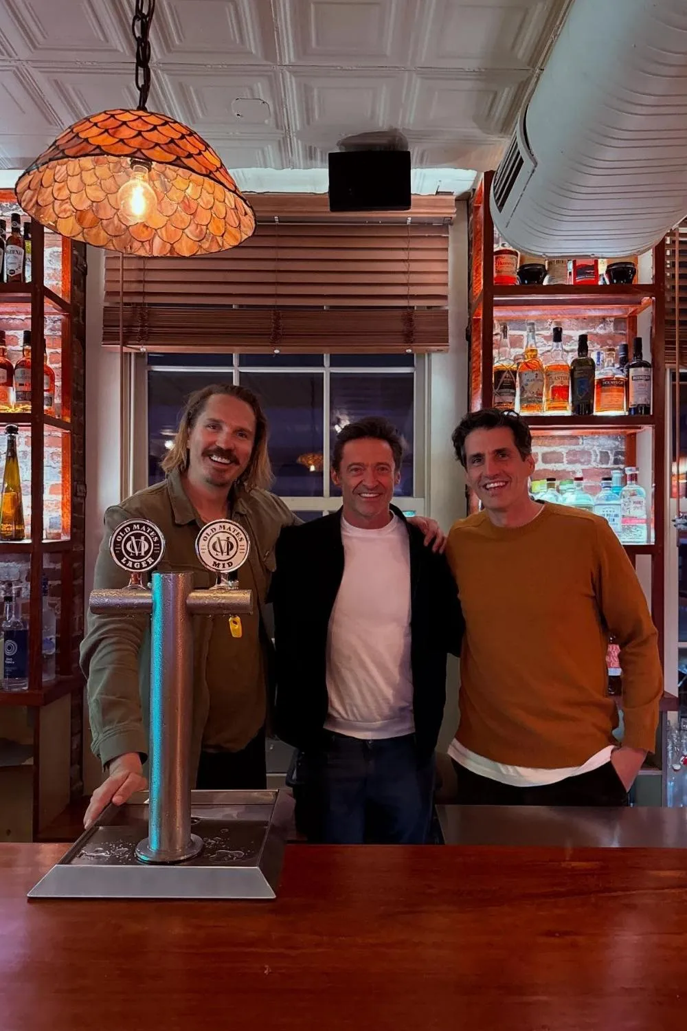 Andy Lee stands behind the bar with Hugh Jackman and another man at Old Mates, the Aussie-owned pub in New York, smiling together in front of shelves stocked with liquor bottles and beer taps.