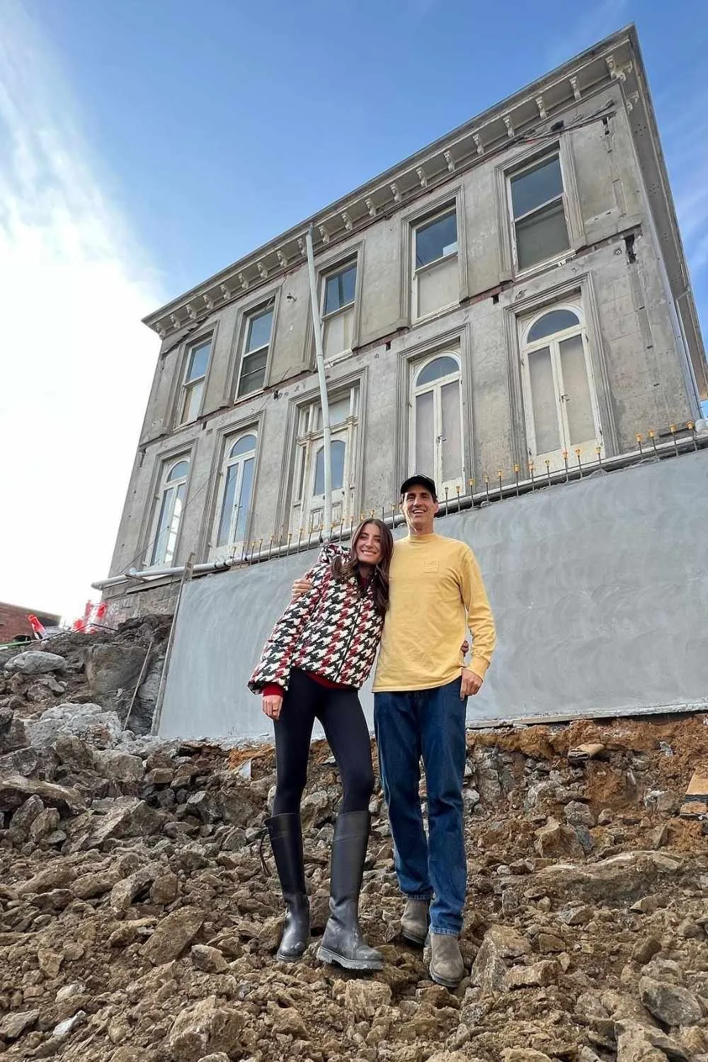 Andy and Bec stand arm-in-arm on a rocky construction site in front of their Melbourne heritage home, during renovation, smiling at the camera.