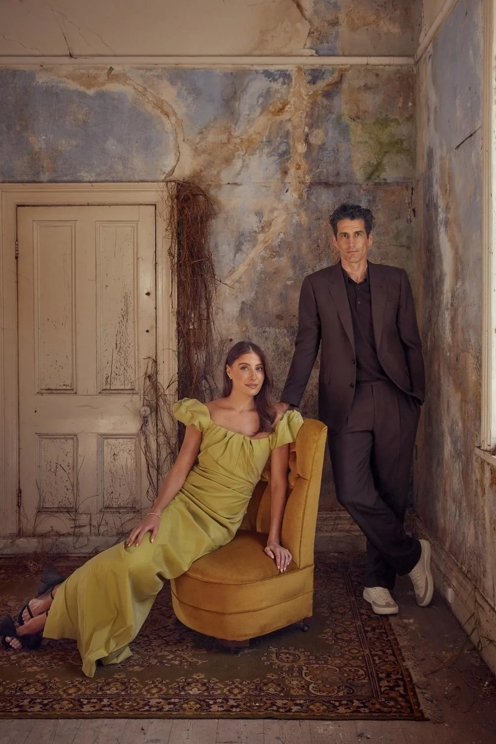 Andy Lee and fiancée Rebecca Harding pose in a moody, rustic room for a photo shoot, dressed in elegant formalwear with vintage textures and earthy tones.