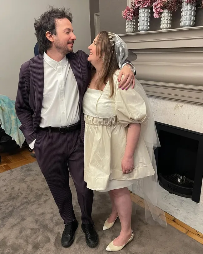 lena dunham and husband with arms around each other at their second wedding with lena in a white cotton mini dress with puffed sleeves