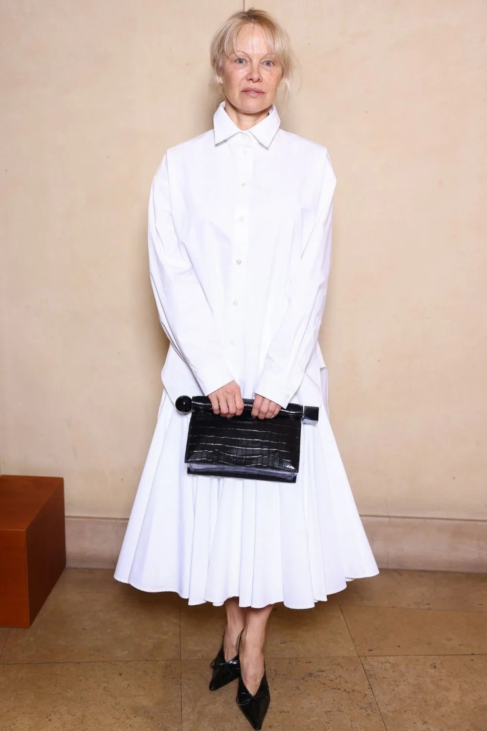 Pamela Anderson style wearing a white collared shirt and pleated white maxi skirt.